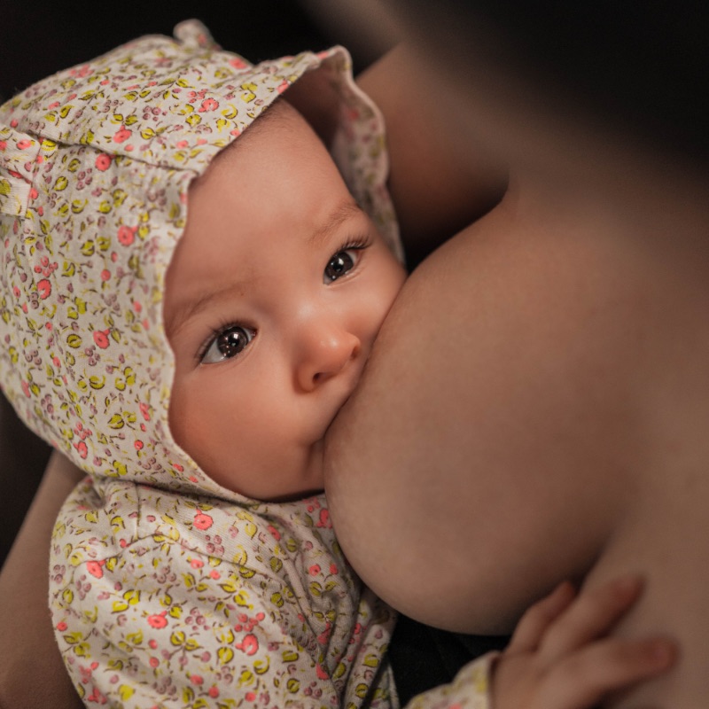 Bild eines braunäugigen Babys im Blümchenjumper, welches gerade an der Brust der Mutter saugt, beziehungsweise gestillt wird.