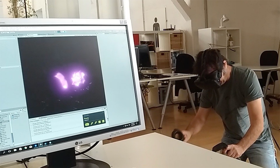 Man sieht im Vordergrund einen Computerbildschirm mit dem Protototypen des Spiels „The Tear“, bei dem gerade zwei Feen-Kristalle zusammengefügt werden. Im Hintergrund sieht man den Gamedesigner Lev Lumesberger mit VR-Brille und Controllern, wie er das Spiel testet.