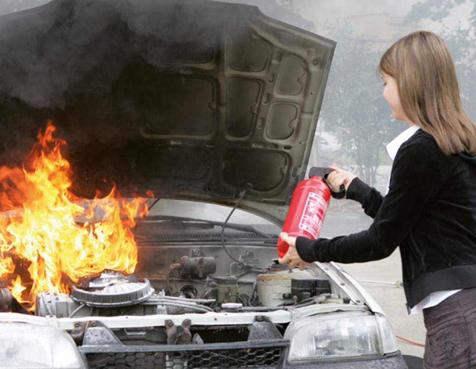 Eine Frau richtet einen kleinen Feuerlöscher auf den offenen, brennenden Motorraum eines PKW.