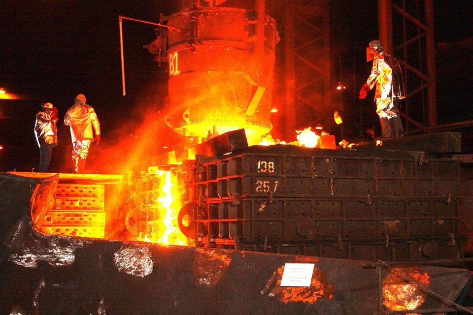 Stahlschmelze in einem Stahlwerk, mit grell in gelb bis rot Tönen glühendem Stahl in der Mitte und drei Männern in Schutzmontur, Arbeitskleidung.