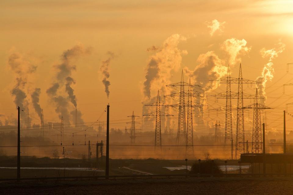 Industriezone mit vielen Strommasten und rauchenden Schornsteinen, eingehüllt in tief orangenem Sonnenlicht.