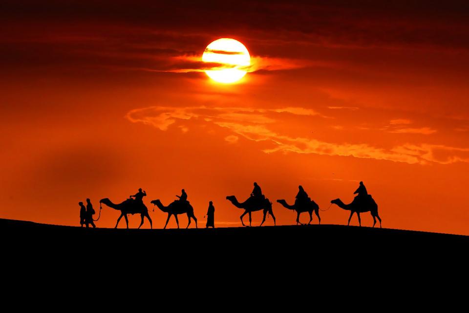 Eine Karavane zieht im Abendrot über eine Düne in der Wüste, von den Kamelen und deren Treibern ist vor dem roten Himmel nur noch die Sillhouette erkenntlich. Die Düne ist schwarz abgebildet. 