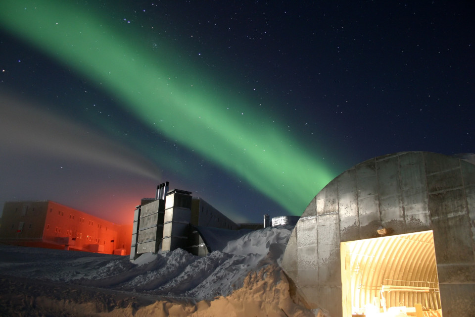 Ein Bild einer Forschungsstation in der Antarktis mit grün schimmernden Polarlichtern am Sternenhimmel.