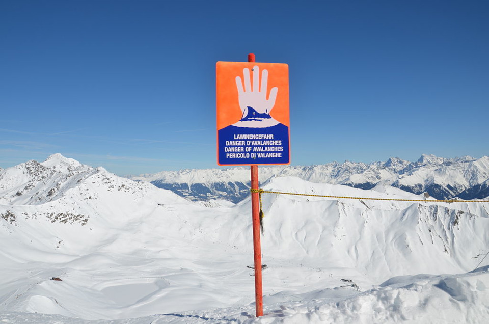 Lawinenwarnschild auf einer schneebedekten Fläche, das Schild ist neon-orange, zur Hälfte mit einer weißen Hand bedruckt.