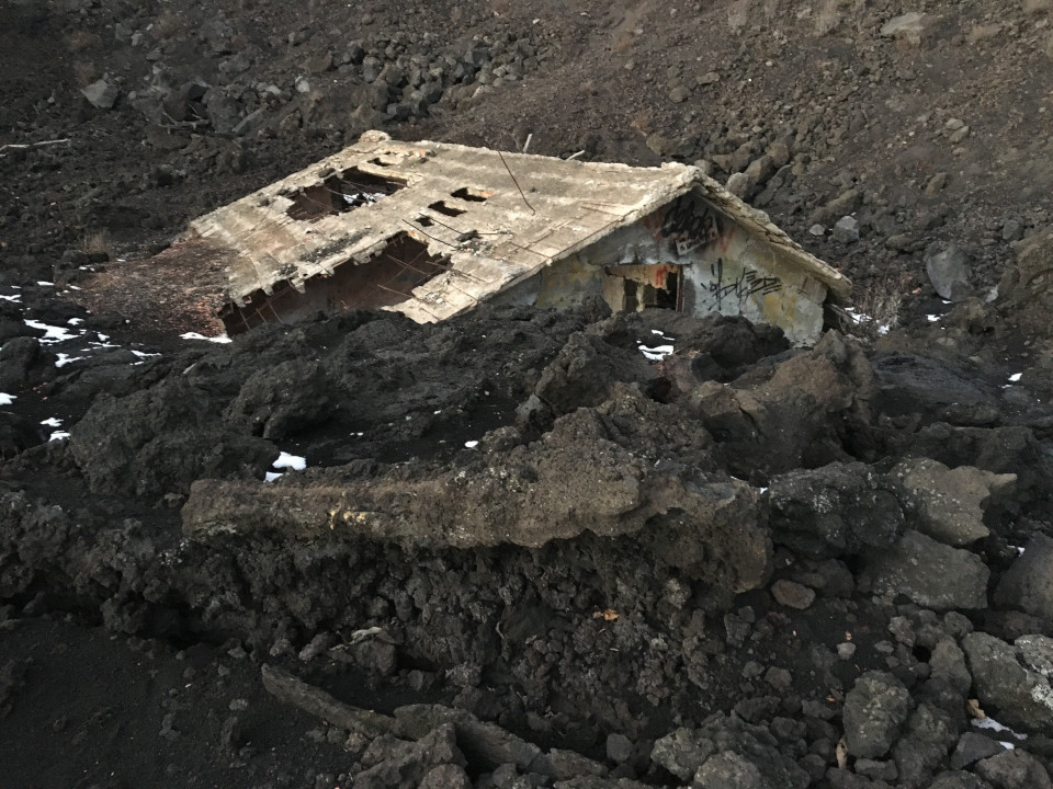 Ein Haus, dessen Dach sichtbar ist, der Rest wurde von Lava zerstört und liegt nun in schwarzem Vulkagestein begraben. 