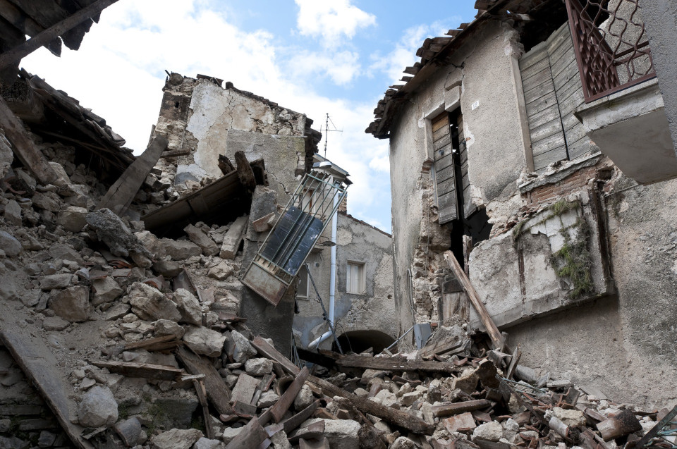 Bild direkt aus einem Trümmerfeld nach einem Erdbeben, rundherum zerstörte Gebäude, graue Steinsbrocken.