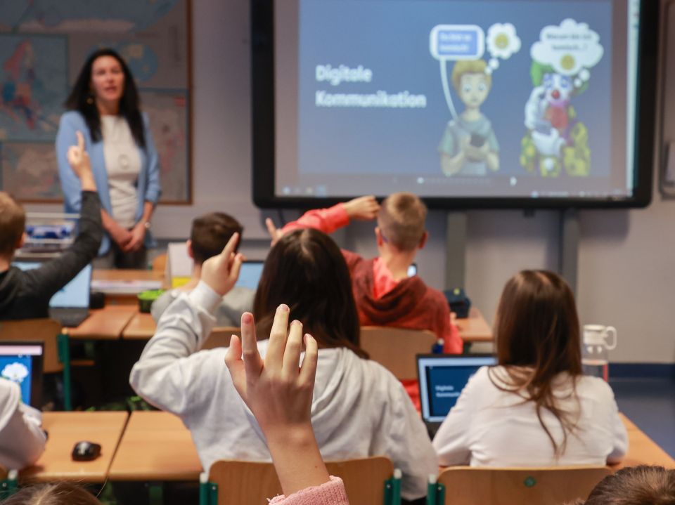 Unterrichtssituation: Eine Lehrerin unterrichtet vor einer digitalen Tafel, die SchuBu+ zeigt. Im Vordergrund sind ihre Schüler*innen zu sehen, die mitarbeiten.